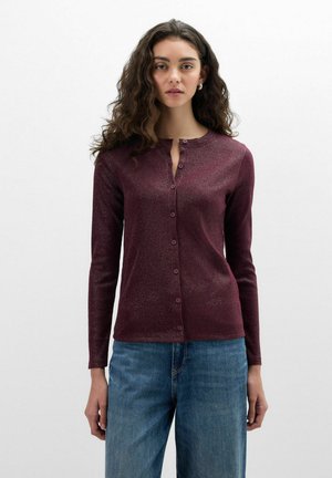 Frau mit langen lockigen Haaren, die ein bordeauxfarbenes, langärmeliges Oberteil mit Knöpfen und blaue Jeans trägt, steht vor einem schlichten hellen Hintergrund.