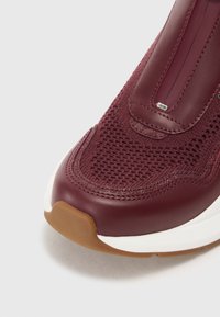 Maroon sneaker with a textured mesh upper and smooth leather accents. Features a rounded toe and a white midsole with a brown rubber outsole.