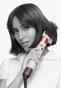 Hårtorkborste med en rosa och vit design, utrustad med ett borsthuvud och ett strukturerat grepp. Användaren har hår av medellängd och mörk färg.