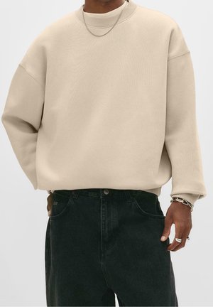 Mann trägt beigen übergroßen Pullover, dunkle Jeans, silberne Kettenkette, Armband und Ringe, Hand liegt nahe der Tasche.