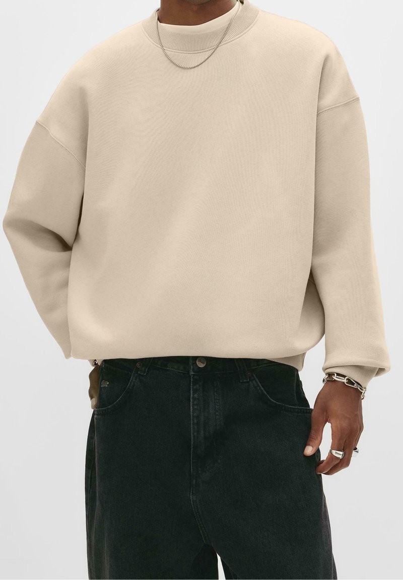 Homme portant un pull beige oversize, un jean foncé, un collier chaîne argenté, un bracelet et des bagues, la main reposant près de la poche.