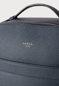 Tamsiai mėlynas odinis kuprinės modelis su tekstūruotu paviršiumi, turintis viršutinį rankeną, užtrauktuko uždarymą ir iškilų "LANCEL PARIS" logotipą.