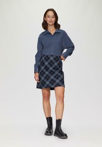 Chemise bleue à manches longues, assortie à une jupe à carreaux bleu marine avec des lignes bleu clair, et des bottines noires.