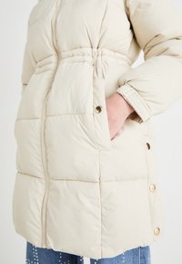 Abrigo acolchado beige con cintura ajustada, con cierre de cremallera, bolsillos laterales y botones de presión en tono dorado a lo largo del dobladillo. Textura suave y acolchada.