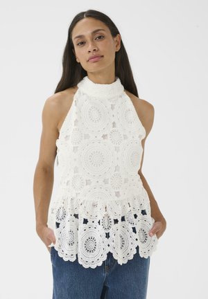 Vrouw draagt een mouwloze witte gehaakte kanten top met hoge hals en gerimpelde zoom, gecombineerd met blauwe spijkerbroek, staand met handen in de zakken.