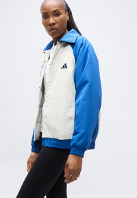 Γυναίκα που φοράει μπλε και λευκό μπουφάν Adidas και μαύρο κολάν, στέκεται μπροστά σε ένα απλό φωτεινό φόντο.