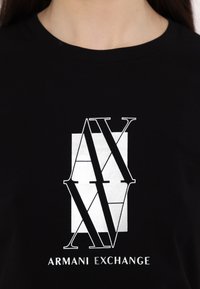 Svart T-skjorte med et stort hvitt grafisk motiv med bokstavene "AX" og "Armani Exchange" i fet skrift. Glatt bomullsstoff.