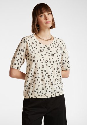 Suéter de punto beige de manga corta con un patrón floral negro, que presenta un cuello redondo y un corte relajado, combinado con pantalones negros.
