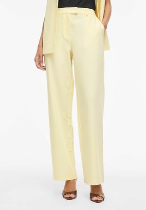 Nő világos sárga, magasderekú, egyenes szárú nadrágot visel oldalzsebekkel, barna nyitott orrú magassarkú szandálokkal párosítva.