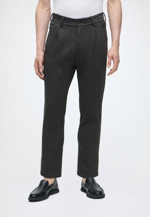 Hombre con pantalones de vestir ajustados gris oscuro, zapatos negros sin cordones y camisa blanca, de pie frente a un fondo claro y liso.