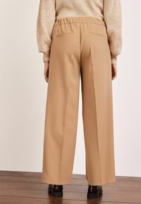 Pantalons larges beiges à taille haute élastique, avec deux poches arrière et des lignes épurées. Fabriqués dans un tissu lisse avec une coupe ajustée.