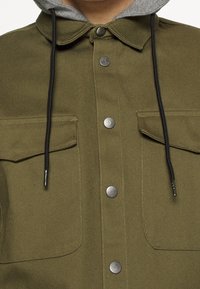 Chaqueta verde oliva con bolsillos de solapa en el pecho, botones a presión plateados en el frente y una capucha gris con cordones negros.