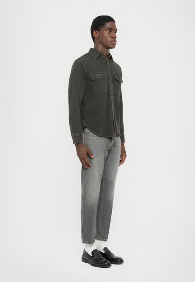 Dunkelgraues Hemd mit zwei Brusttaschen, kombiniert mit hellgrauen Jeans. Legere schwarze Slipper und weiße Socken runden das Outfit ab.