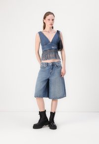 Top corto in denim con dettagli in frange, abbinato a shorts in denim lunghi fino al ginocchio. Il modello indossa stivali da combattimento neri con lacci. Borsa nera semplice.
