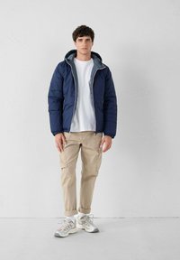 Joven de pie con chaqueta azul marino con capucha, camiseta blanca, pantalones cargo beige, calcetines blancos y zapatillas grises sobre fondo blanco liso.
