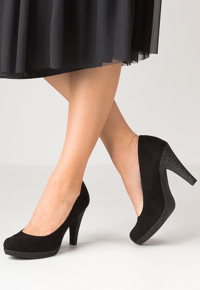 Femme portant des escarpins en daim noir avec des talons scintillants et une jupe noire, jambes croisées sur un fond clair uni.