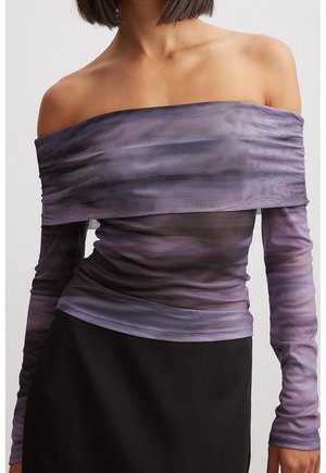 Vrouw draagt een doorzichtige paarse off-shoulder top met lange mouwen, gecombineerd met een zwarte rok, waarbij de halslijn en de bovenlichaam zichtbaar zijn.