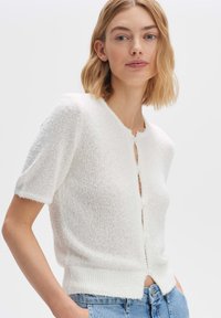 Weißer Strickcardigan mit kurzen Ärmeln, strukturiertem Material, rundem Ausschnitt und Knopfverschluss. Weiche Oberfläche mit rohen Kanten.