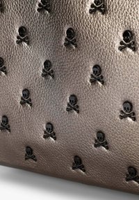 Bolso de cuero bronce metálico que presenta motivos de calaveras y huesos cruzados negros en relieve, distribuidos uniformemente por toda la superficie. Acabado texturizado.