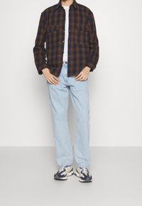 Rutigt brunt och marinblått skjorta med två framfickor, bärs över en vit t-shirt, kombinerat med ljusblå jeans och flerfärgade sneakers.