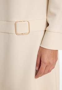 Hand ruht neben einem cremefarbenen Mantel mit einem Stoffgürtel und quadratischem goldfarbenen Schnallen-Detail in der Taille.