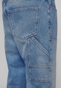 Jeans en denim bleu clair avec un design à cinq poches, une texture délavée et des coutures contrastantes. Poche arrière inclinée avec des accents rivetés.