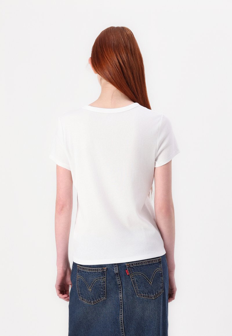 Weißes T-Shirt mit kurzen Ärmeln und rundem Halsausschnitt, getragen mit einem Jeansrock, der zwei Gesäßtaschen und ein rotes Tab-Detail aufweist.