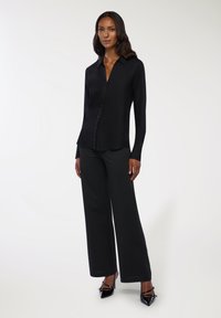 Czarna koszula zapinana na guziki z gładkiego materiału, z kołnierzykiem, zestawiona z szerokimi spodniami w paski. Obuwie to czarne czółenka z wydłużonym noskiem.