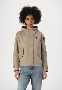 Giacca beige con cappuccio, chiusura con bottoni frontale, due tasche anteriori e una toppa sulla manica. Tessuto misto cotone con una texture liscia.