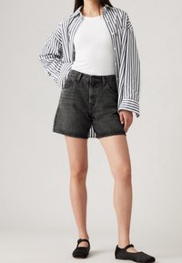 Pantalones cortos de mezclilla negros con dobladillos desgastados, combinados con una camiseta sin mangas blanca y una camisa de rayas en blanco y negro de corte holgado. Zapatos negros sin cordones.