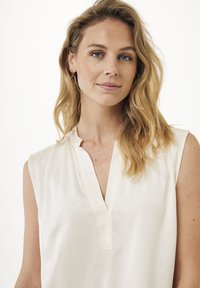 Ärmlös, krämfärgad blus med V-ringning och subtil lyster. Mjuk tygkvalitet med minimalt detaljer, slät textur och avslappnad passform.