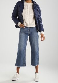 Granatowa kurtka puchowa z kapturem, kremowy sweter z dzianiny z wzorem w warkocze, krótkie dżinsy z postrzępionymi brzegami i białe trampki.