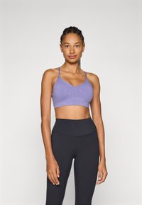 Sutiã desportivo lavanda com alças finas e design em V. O tecido apresenta uma textura subtil. Combinado com leggings pretas de cintura alta.