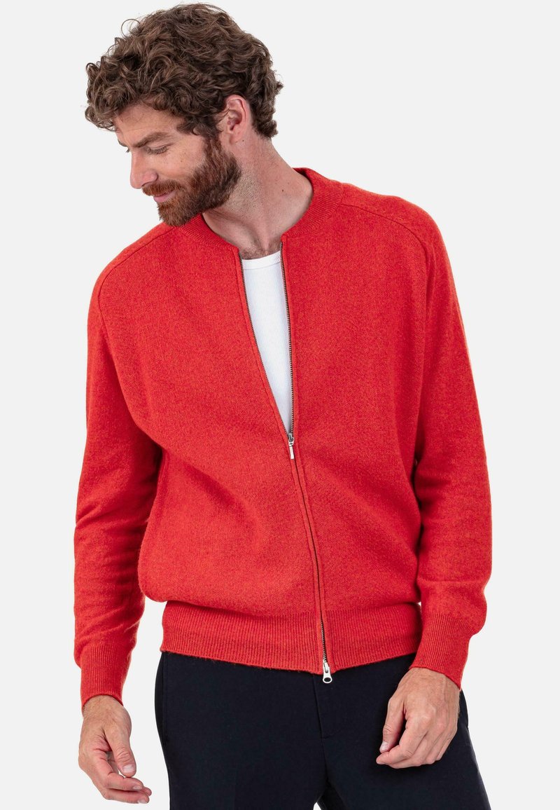 Roter Pullover mit Reißverschluss, rundem Ausschnitt, gerippten Bündchen und Saum. Weiche Textur, figurnah geschnitten, über einem weißen Hemd getragen.