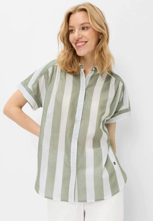 Femme souriante aux cheveux blonds portant une chemise à manches courtes à larges rayures verticales vertes et blanches, debout, la main sur la hanche.