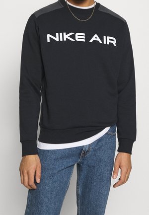 Osoba nosząca czarną bluzę Nike Air na białej koszuli i niebieskich dżinsach, widoczny cienki złoty łańcuszek.