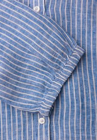 Manche de chemise rayée verticalement bleu et blanc avec poignet élastique et boutons blancs sur un fond en tissu rayé assorti.