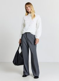 Pull en laine blanc avec des embellissements texturés, associé à un pantalon large à rayures grises. Le mannequin tient un sac à main noir décontracté.