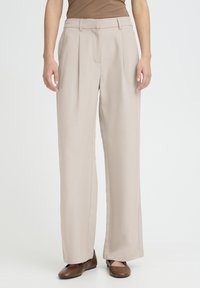 Pantalon beige à jambes larges avec plis à l'avant et poches latérales, en tissu lisse, associé à des chaussures plates marron. Coupe ajustée et décontractée.