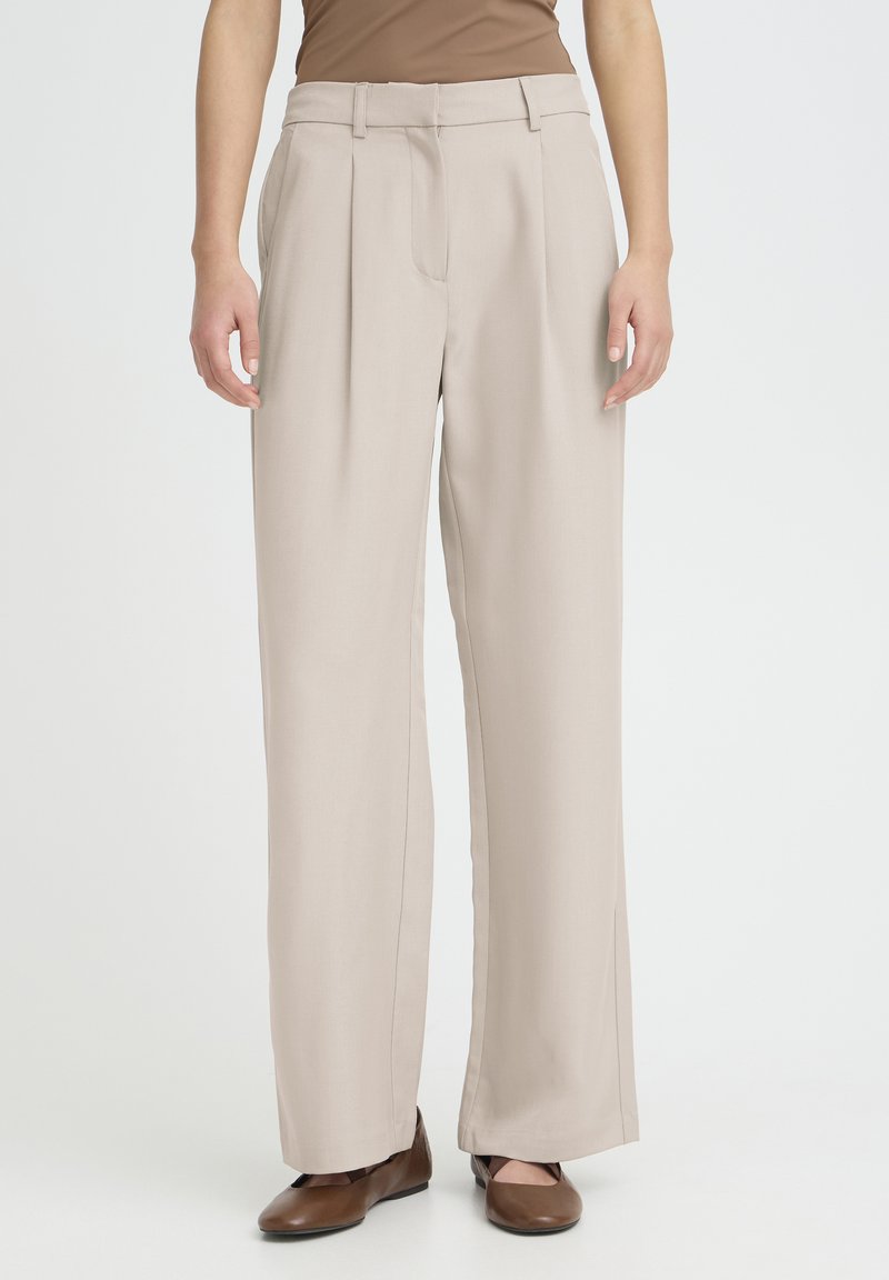 Pantalon beige à jambes larges avec plis à l'avant et poches latérales, en tissu lisse, associé à des chaussures plates marron. Coupe ajustée et décontractée.