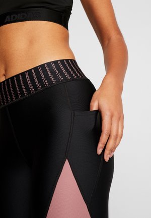 Zbliżenie osoby noszącej czarne i różowe legginsy z boczną kieszenią, ręką spoczywającą w kieszeni, widoczny stanik sportowy Adidas.