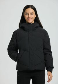 Frau mit langen dunklen Haaren, die eine schwarze Daunenjacke mit hohem Kragen trägt, steht mit einer Hand in der Tasche und lächelt in die Kamera.