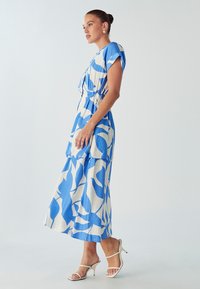 Plava i bijela haljina s uzorkom, kratkim rukavima, strukirana u struku i lepršava suknja. Nosena s bijelim sandalama s trakama. Glatka tekstura tkanine.