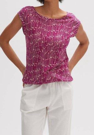 Vrouw draagt een paarse, kortemouwen top met patroon en witte broek, staat met handen in de zij tegen een effen achtergrond.