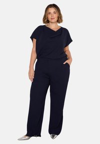 Mörkblå topp med draperad urringning, tillsammans med matchande vida byxor; silverfärgade spetsiga skor ger en kontrasterande detalj.