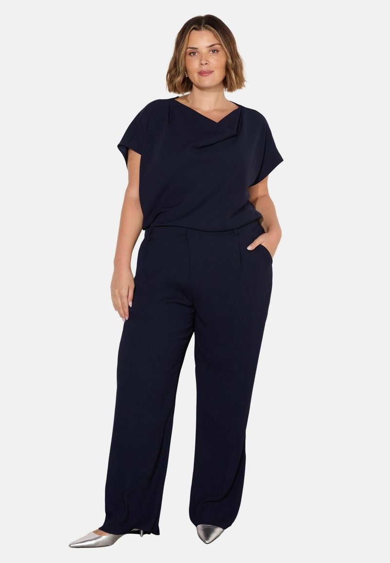 Mörkblå topp med draperad urringning, tillsammans med matchande vida byxor; silverfärgade spetsiga skor ger en kontrasterande detalj.