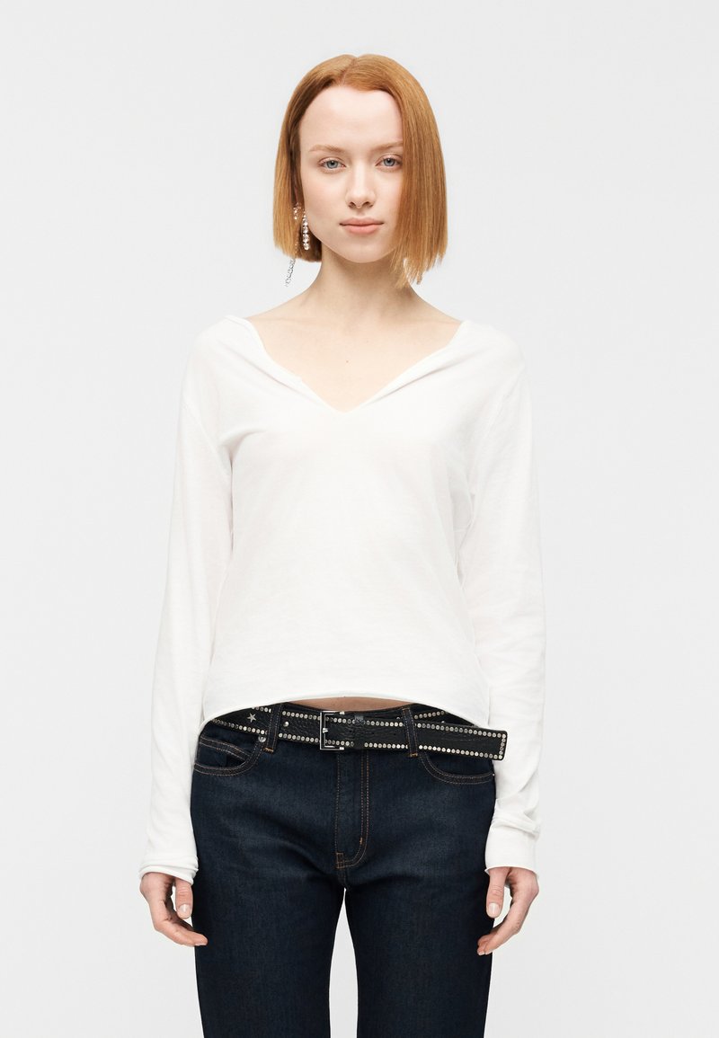 Λευκό μακρυμάνικο τοπ με V-λαιμόκοψη, σε συνδυασμ ό με σκούρο μπλε τζιν και μαύρη ζώνη με τρουκς. Το ύφασμα φαίνεται μαλακό και η εφαρμογή άνετη.