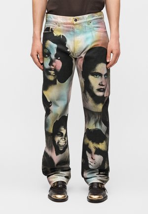 Męskie wielokolorowe jeansy z dużymi wyblakłymi czarnymi i pastelowymi portretami różnych twarzy, w połączeniu z czarnymi mokasynami ze złotymi końcówkami.