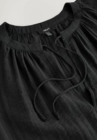 Schwarze Bluse mit rundem Ausschnitt, Raffung und einer vorderen Schleife mit Perlenakzenten. Hergestellt aus strukturiertem, leichtem Stoff.