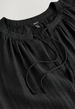 Black blouse with a round neckline, gathered detailing, and a front tie featuring bead accents. Made of textured, lightweight fabric.